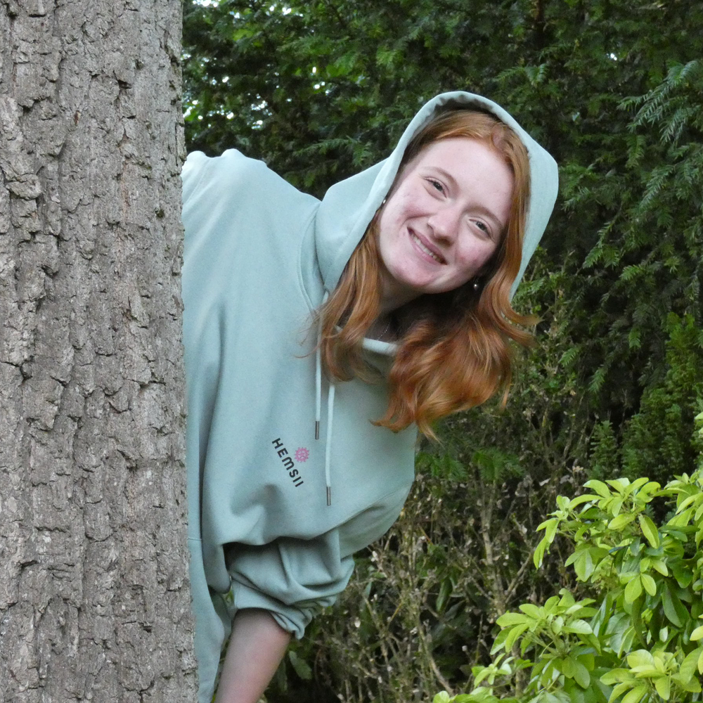 Person wearing an aloe hoodie peeking from behind a tree in a green outdoor setting